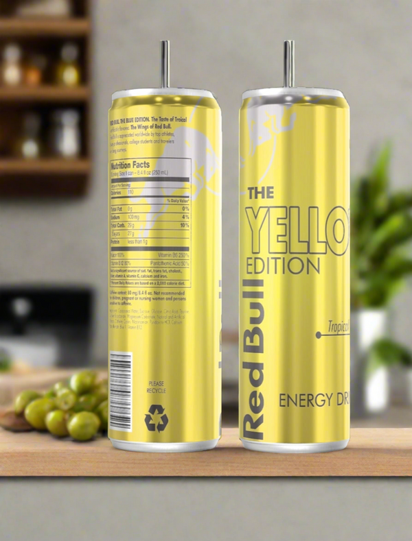 A Yellow Tropical Red Bull 20oz Tumbler by Kreative Kreationz sits on a wooden surface beside two yellow Red Bull cans—one labeled "The Yellow Edition," the other showing nutrition facts—with a blurred kitchen background.