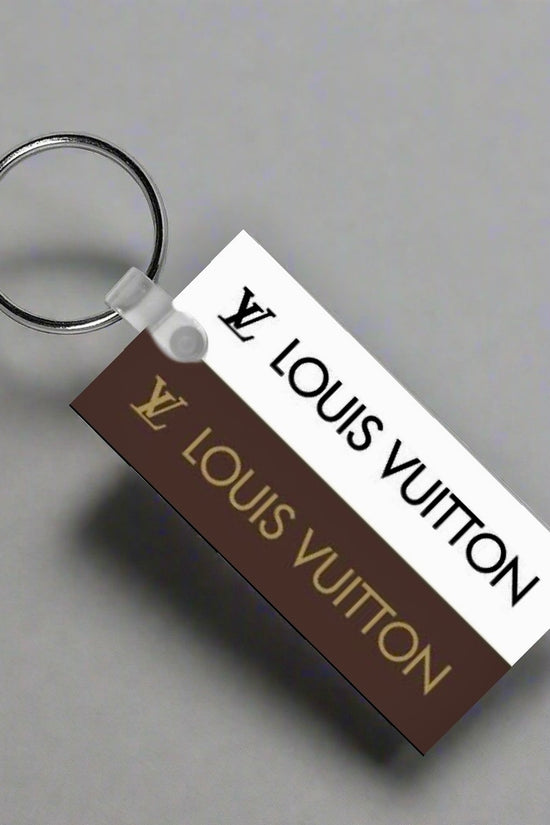 The Louis V Keychain by Kreative Kreationz features a durable rectangular tag with the Louis Vuitton logo and name in gold on brown and black on white—an elegant accessory for any collection.