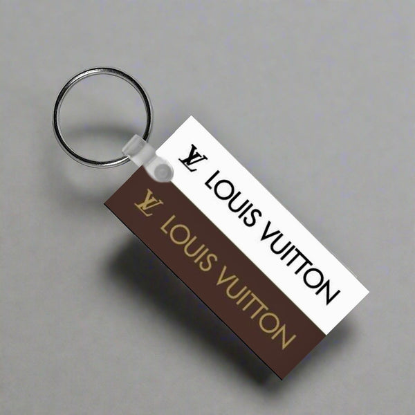 The Louis V Keychain by Kreative Kreationz features a durable rectangular tag with the Louis Vuitton logo and name in gold on brown and black on white—an elegant accessory for any collection.