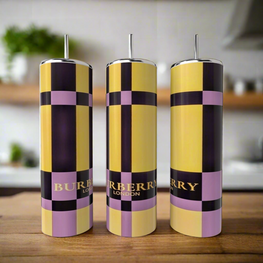 Three tall, cylindrical Kreative Kreationz Burberry 20oz Skinny Tumblers with metal straws sit on a wooden surface. The stainless steel set features yellow, black, and lavender checks with "BURBERRY LONDON" in gold; a blurred kitchen is visible.