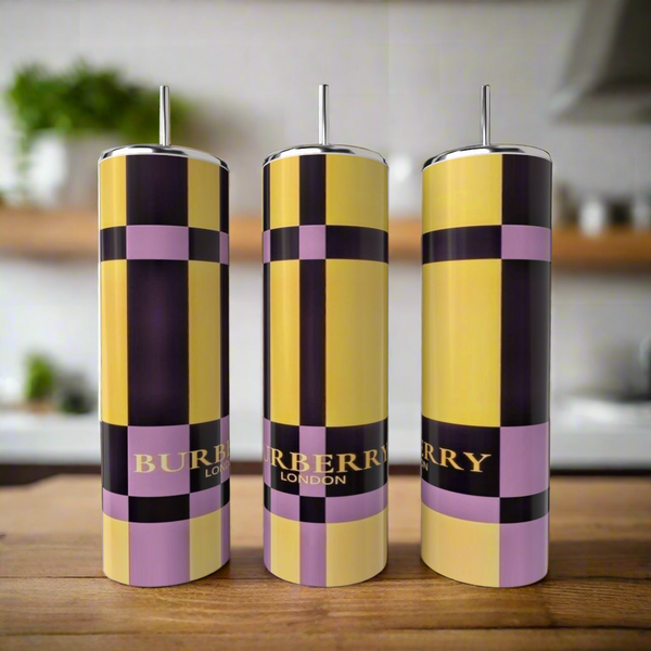 Three tall, cylindrical Kreative Kreationz Burberry 20oz Skinny Tumblers with metal straws sit on a wooden surface. The stainless steel set features yellow, black, and lavender checks with "BURBERRY LONDON" in gold; a blurred kitchen is visible.