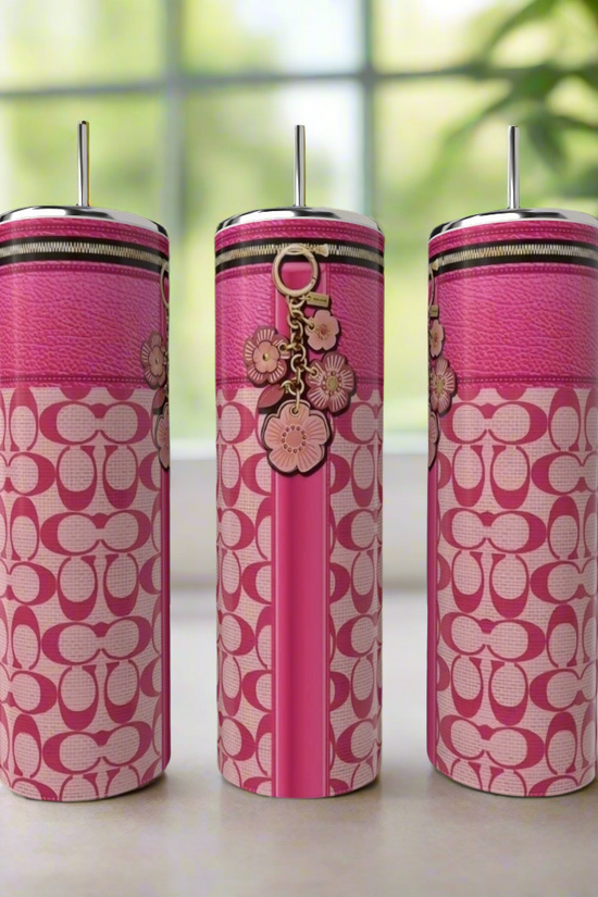 Three tall, pink Coach 20oz Tumblers by Kreative Kreationz, each with a silver straw, patterned design, and floral charm, are displayed on a light surface with a blurred window and green plant in the background.