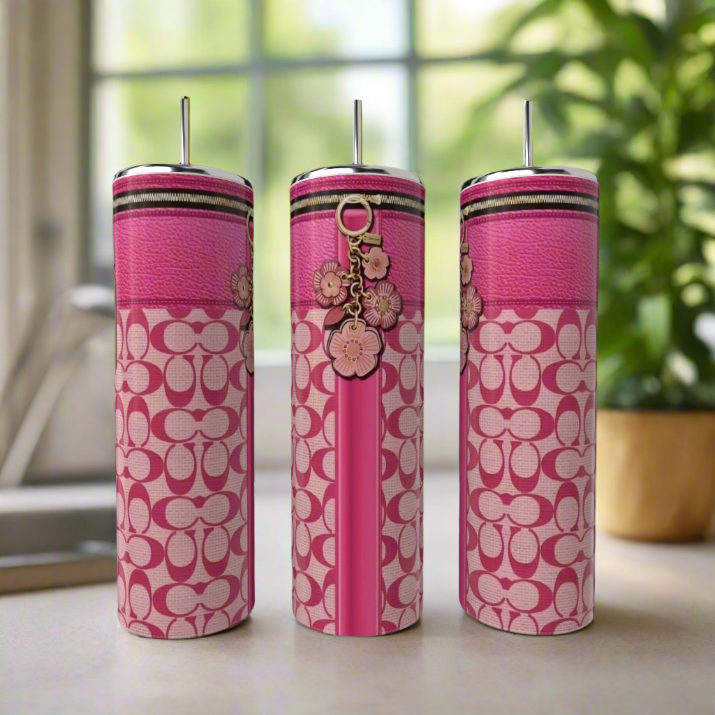Three tall, pink Coach 20oz Tumblers by Kreative Kreationz, each with a silver straw, patterned design, and floral charm, are displayed on a light surface with a blurred window and green plant in the background.