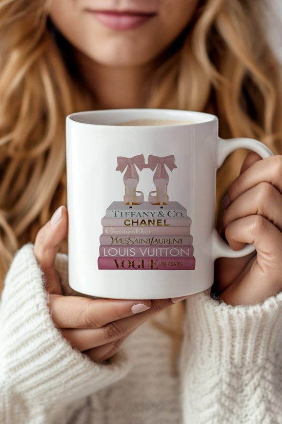 A woman in a cozy white sweater holds the Kreative Kreationz Designer Inspired 11oz Ceramic Mug, featuring stacked fashion books labeled Tiffany & Co., Chanel, YSL, Louis Vuitton, and Vogue with a pink bow on top.