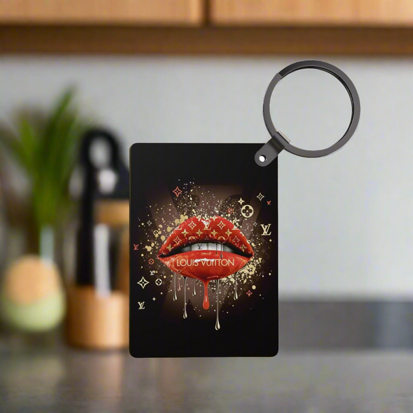 The Drip Lips LV Inspired Keychain by Kreative Kreationz features glossy red lips with gold drip and luxe accents, displayed in focus against a blurred kitchen backdrop.