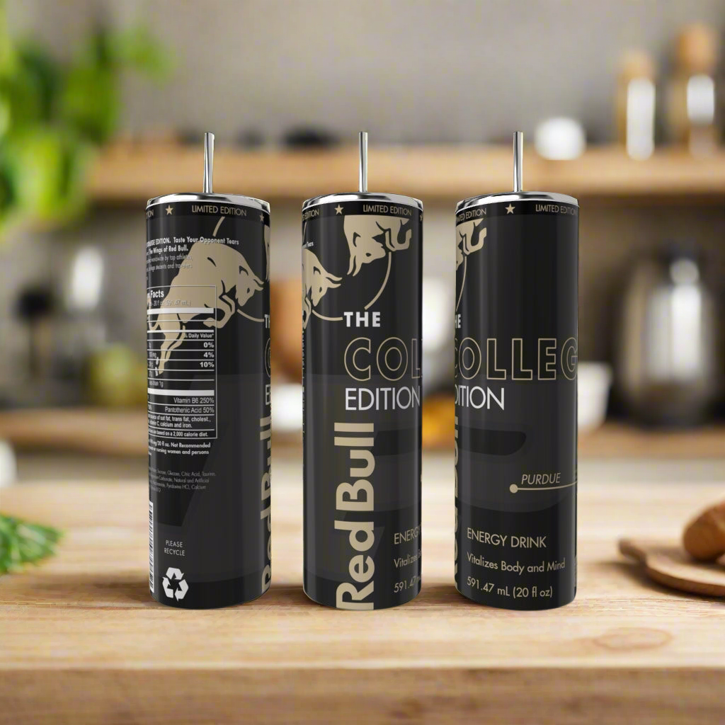 Three limited edition Red Bull cans labeled "The College Edition Purdue" stand on a wooden kitchen counter next to a Kreative Kreationz College Edition 20oz Tumbler, with a blurred background of kitchen items and greenery.