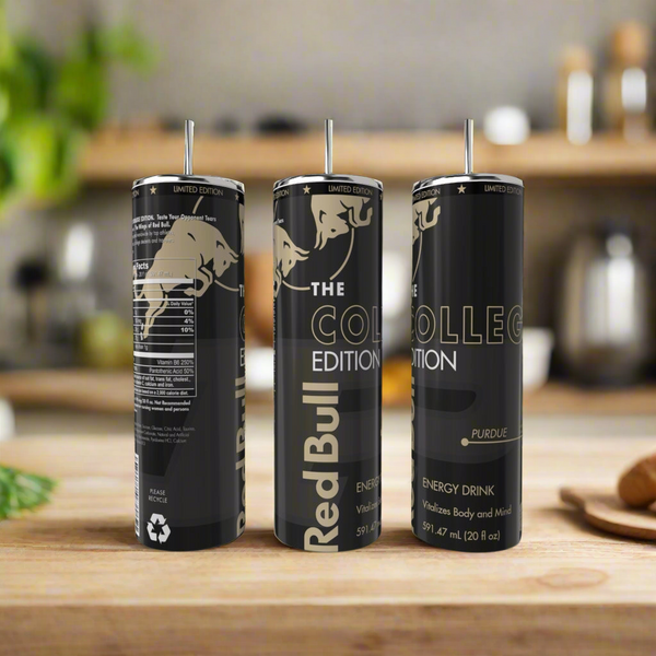 Three limited edition Red Bull cans labeled "The College Edition Purdue" stand on a wooden kitchen counter next to a Kreative Kreationz College Edition 20oz Tumbler, with a blurred background of kitchen items and greenery.