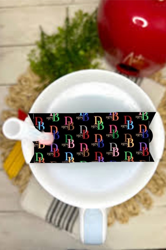 A white teacup and saucer with a spoon sit on a round mat, topped with the Dooney & Bourke Stanley Lid Plate/Topper by Kreative Kreationz. A black strip with colorful “RB 1975” text adds flair, with a red teapot and green plant in the background.