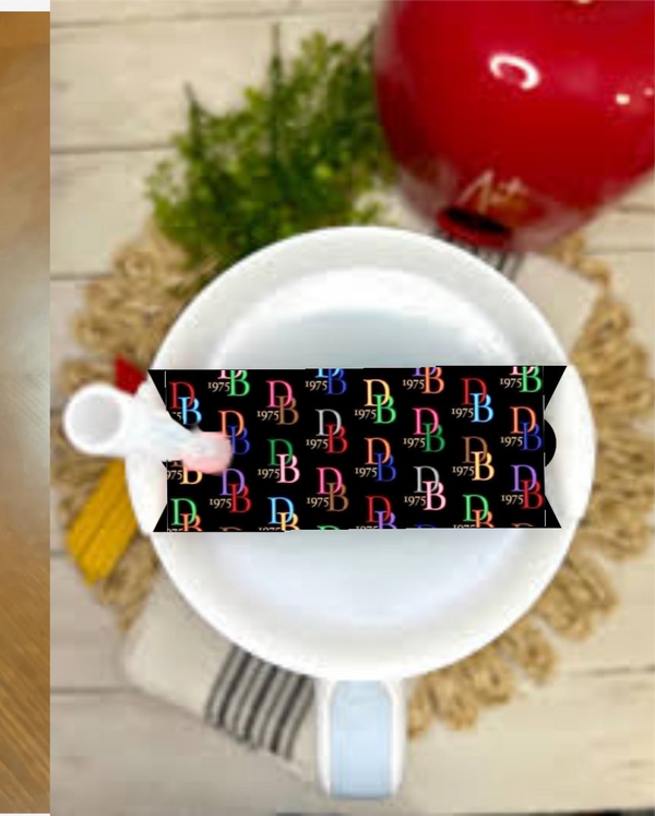 A white teacup and saucer with a spoon sit on a round mat, topped with the Dooney & Bourke Stanley Lid Plate/Topper by Kreative Kreationz. A black strip with colorful “RB 1975” text adds flair, with a red teapot and green plant in the background.