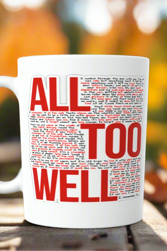 The ALL TOO WELL LYRICS- RED Era-Taylor Swift Ceramic Mug by Kreative Kreationz, featuring bold red and black text, rests on a wooden outdoor table amid blurred autumn trees with orange leaves in the background.