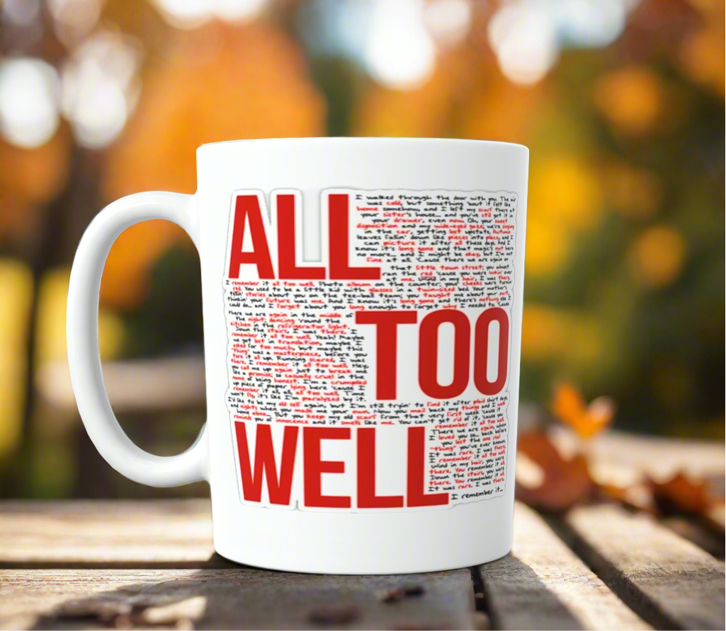 The ALL TOO WELL LYRICS- RED Era-Taylor Swift Ceramic Mug by Kreative Kreationz, featuring bold red and black text, rests on a wooden outdoor table amid blurred autumn trees with orange leaves in the background.