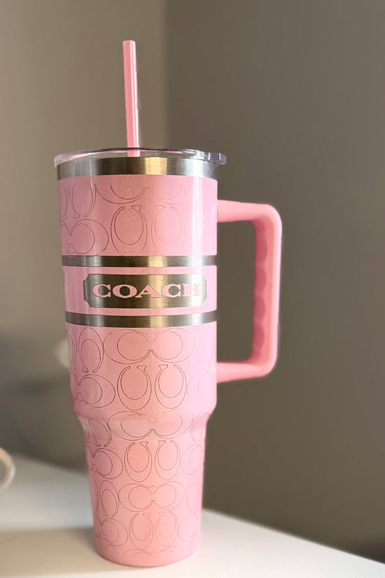 The Coach Pink 40oz Engraved Tumbler by Kreative Kreationz is a large pink stainless steel tumbler with a handle, silver lid, pink straw, embossed pattern, and metallic Coach logo label, displayed on a white surface with a gray background.