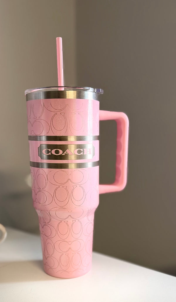 The Coach Pink 40oz Engraved Tumbler by Kreative Kreationz is a large pink stainless steel tumbler with a handle, silver lid, pink straw, embossed pattern, and metallic Coach logo label, displayed on a white surface with a gray background.
