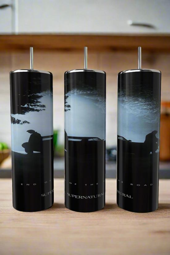The Kreative Kreationz Supernatural 20oz Skinny Tumbler features a luxury car silhouette with two people on its hood under a cloudy sky, crafted from eco-friendly stainless steel and paired with straws. Background: kitchen countertop, blurred objects.