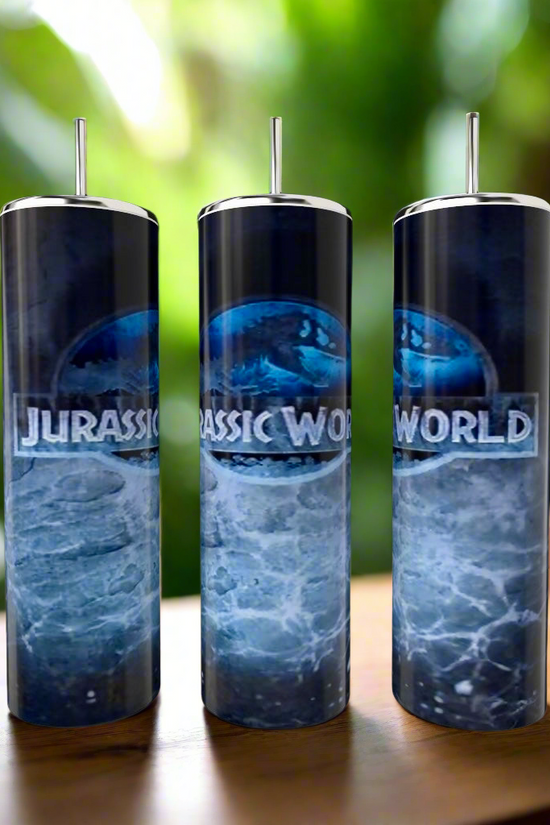 Three elegant Jurassic Park 20oz tumblers by Kreative Kreationz feature luxury-inspired stainless steel with silver lids and are showcased beautifully against ocean waves on a wooden surface, complemented by a blurred green foliage backdrop.