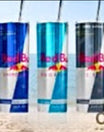 Three Red Bull cans sit on a beach beside a Kreative Kreationz Stainless Steel 20oz Tumbler, which features a spill-proof lid and keeps drinks hot for 4 hours or cold for 12; sun, snowflake, and thermometer icons add flair.