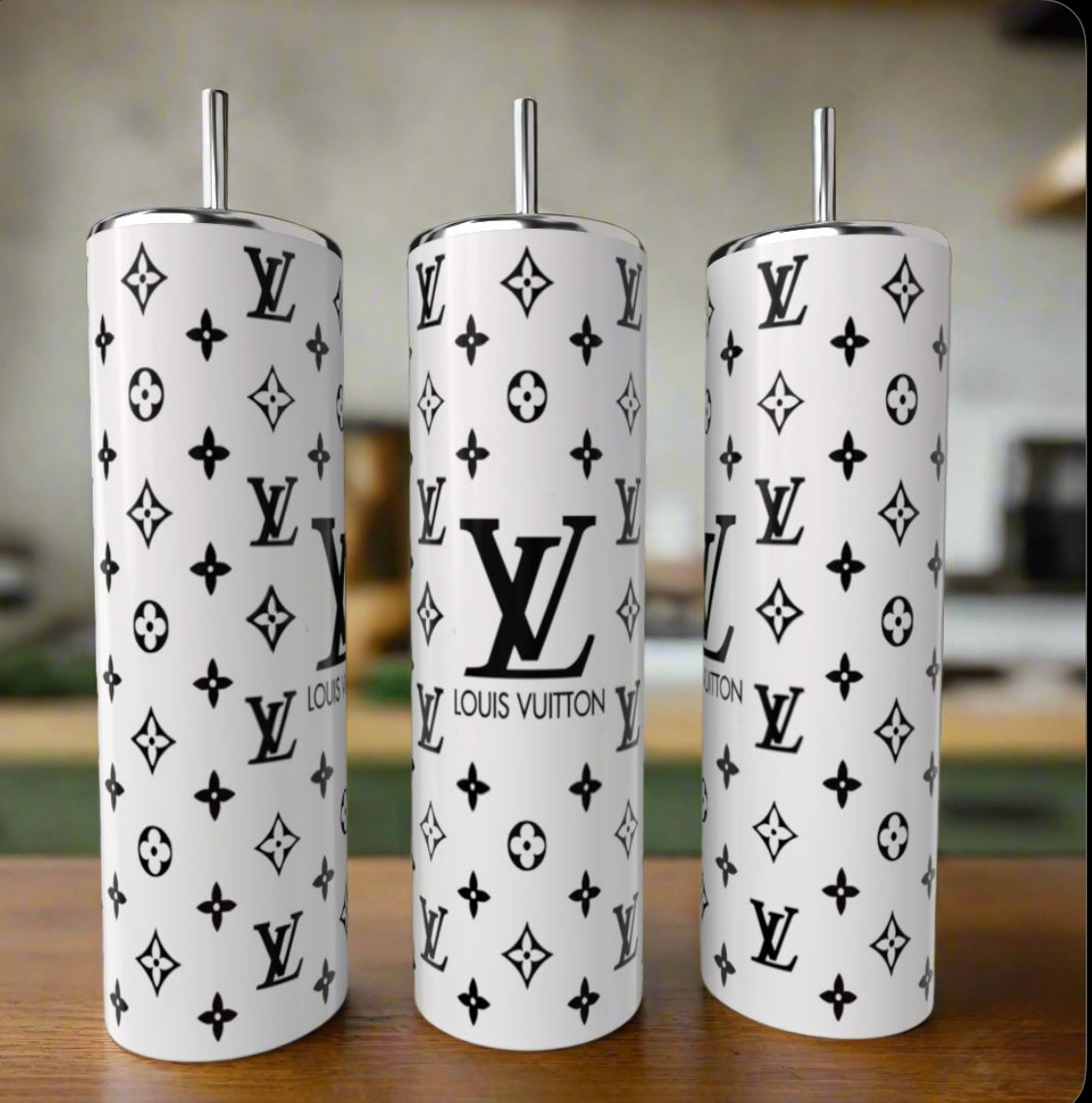 Three tall Kreative Kreationz Black & White LV Basic 20oz tumblers with metal straws rest on wood. Each features the black Louis Vuitton logo and monogram pattern, with a blurred kitchen in the background.
