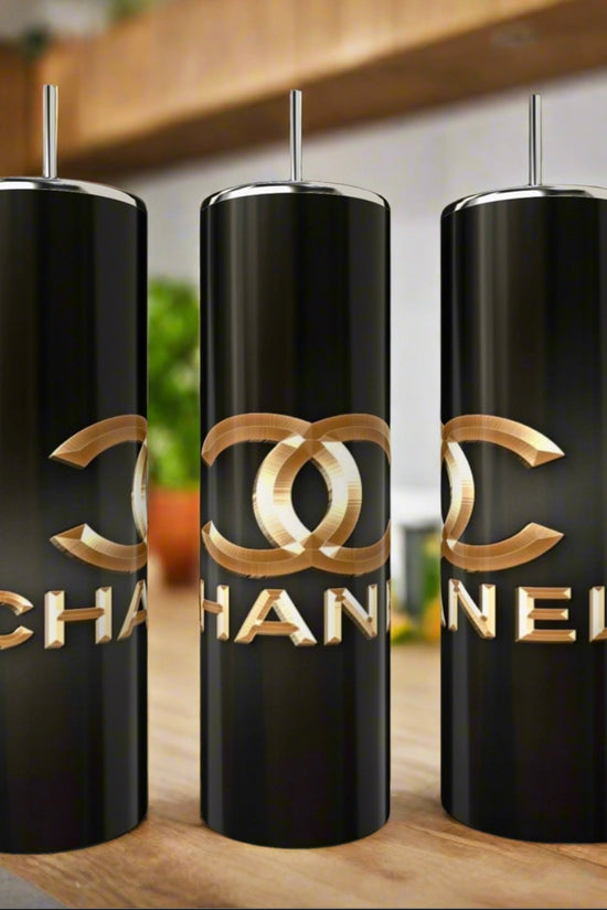 Three Chanel 20oz Skinny Tumblers by Kreative Kreationz, featuring metallic gold logos and text, are displayed on a kitchen counter with blurred plants and shelves in the background.