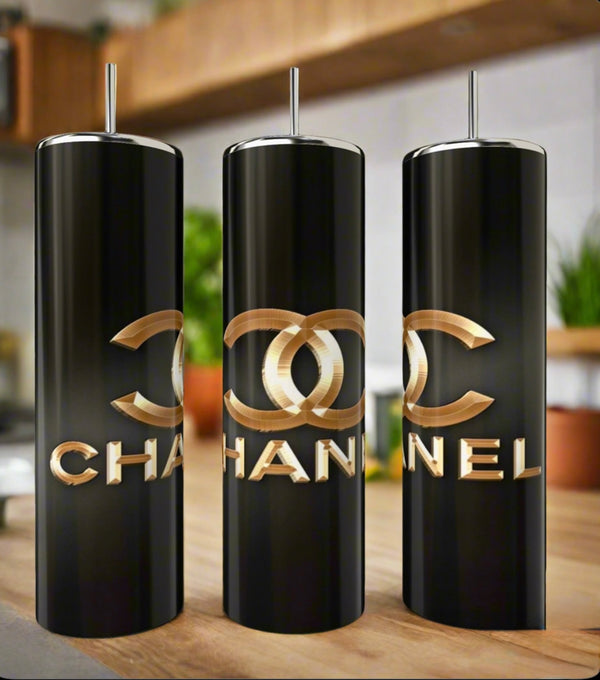 Three Chanel 20oz Skinny Tumblers by Kreative Kreationz, featuring metallic gold logos and text, are displayed on a kitchen counter with blurred plants and shelves in the background.