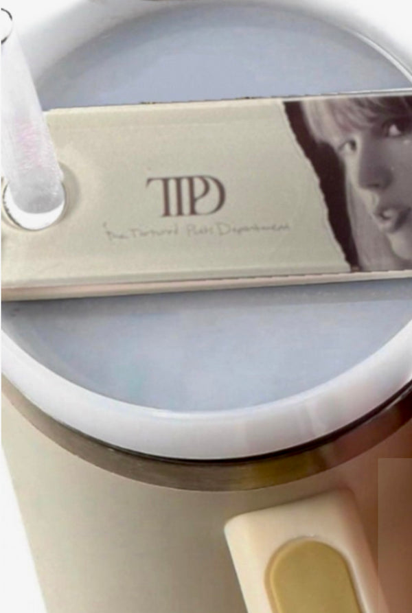 A round, white mug with a frothy drink features the TTPD Taylor Stanley Lid Topper from Kreative Kreationz, resembling a guitar pick with a face and "The Taylor Swift Experience" text. A clear straw is inserted, creating an inviting blend of music and refreshment.