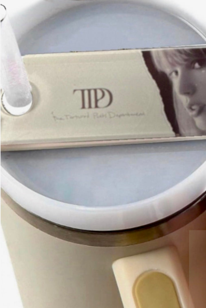 A round, white mug with a frothy drink features the TTPD Taylor Stanley Lid Topper from Kreative Kreationz, resembling a guitar pick with a face and "The Taylor Swift Experience" text. A clear straw is inserted, creating an inviting blend of music and refreshment.