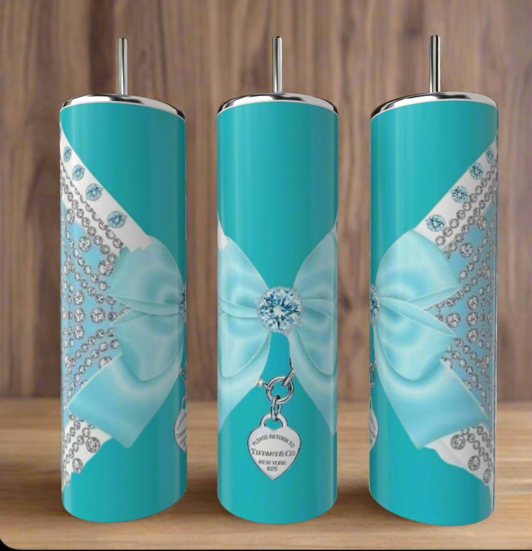 Three Tiffany Lace Locket Wraparound 20oz Tumblers by Kreative Kreationz, featuring silver lids, straws, and jeweled bows with "Return to Tiffany & Co." heart designs, are elegantly displayed on a wooden surface.