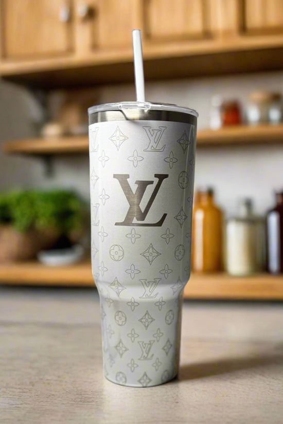 The LV White 40oz Engraved Tumbler by Kreative Kreationz, featuring a repeating Louis Vuitton-inspired pattern and straw, sits on a kitchen counter with shelves and containers blurred in the background.
