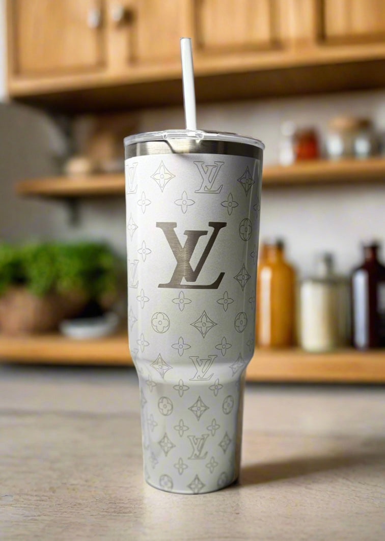 The LV White 40oz Engraved Tumbler by Kreative Kreationz, featuring a repeating Louis Vuitton-inspired pattern and straw, sits on a kitchen counter with shelves and containers blurred in the background.