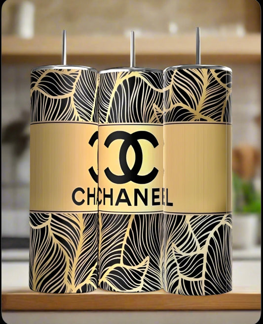 Three 20oz stainless steel tumblers with black and gold leaf patterns are side by side. The center is the "Chanel 20oz Skinny Tumbler | Gold" by Kreative Kreationz, featuring a gold background and logo. A kitchen counter and blurred background appear.