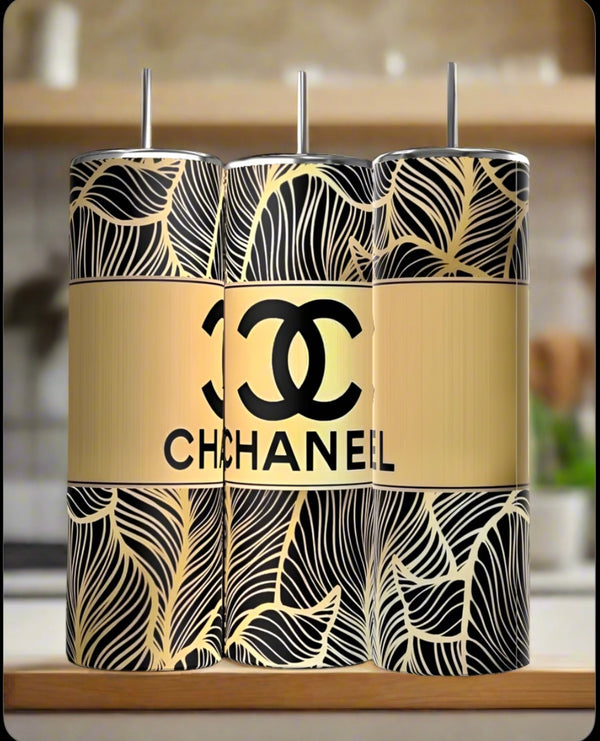 Three 20oz stainless steel tumblers with black and gold leaf patterns are side by side. The center is the "Chanel 20oz Skinny Tumbler | Gold" by Kreative Kreationz, featuring a gold background and logo. A kitchen counter and blurred background appear.