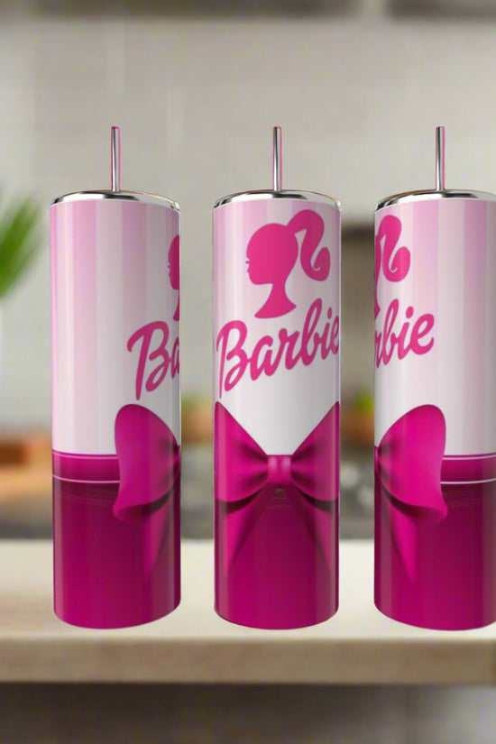 Three Barbie 20oz Tumblers by Kreative Kreationz, each with a pink bow and Barbie logo featuring a woman's silhouette, are lined up on a kitchen counter. The tumblers have straws and the pink-and-white design stands out against a blurred background.