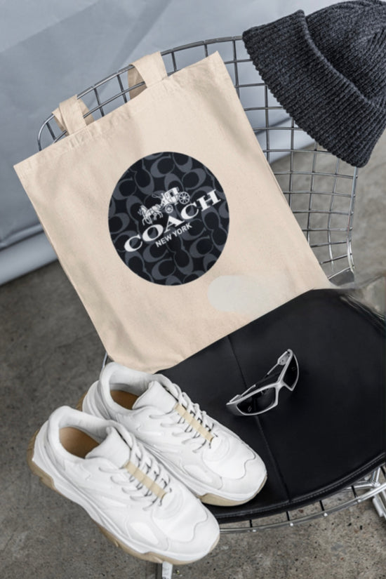 A wire chair displays a COACH Canvas Tote Bag by Kreative Kreationz, featuring a circular logo, surrounded by a gray knit beanie, white sneakers, and sporty sunglasses. The minimalist scene with its concrete floor and gray backdrop may captivate Swifties in search of style inspiration.