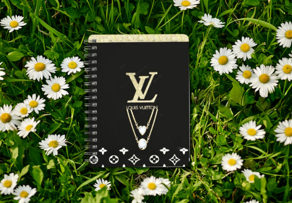 A durable spiral-bound LV A6 Notebook by Kreative Kreationz, featuring a gold "LV" logo and necklace design, rests on grass with white daisies. Adorned with Louis Vuitton motifs at the bottom, it offers elegance and resilience in every page turn.