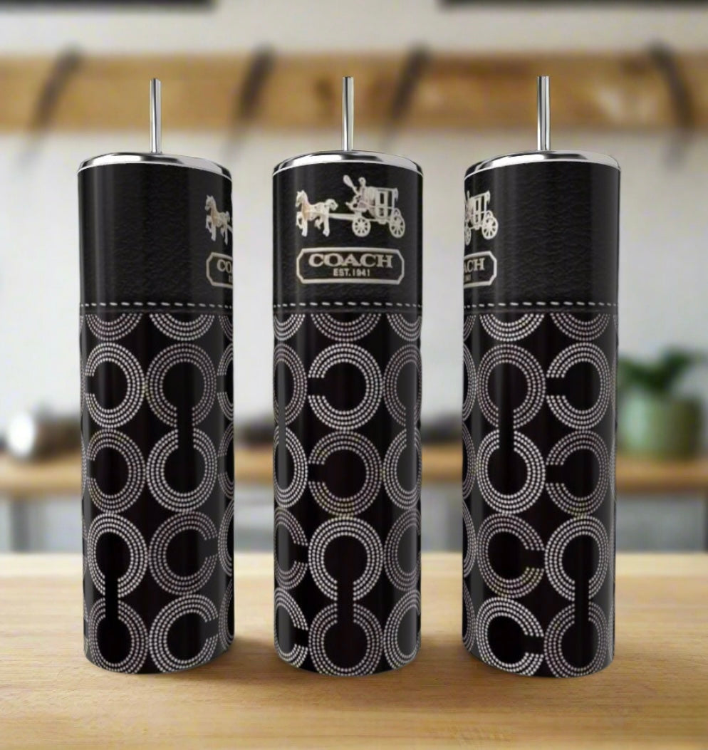Three Kreative Kreationz Coach 20oz Skinny Tumblers, crafted from eco-friendly black metal with silver straws and interlocking circle Coach logos, are displayed on a wooden surface against a blurred kitchen background.