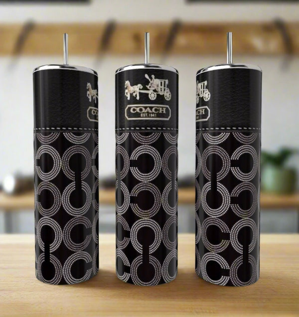 Three Kreative Kreationz Coach 20oz Skinny Tumblers, crafted from eco-friendly black metal with silver straws and interlocking circle Coach logos, are displayed on a wooden surface against a blurred kitchen background.