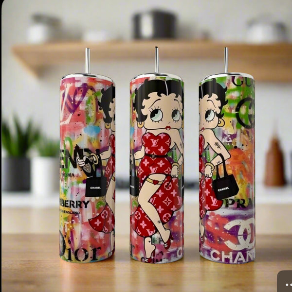 The Kreative Kreationz Betty Boop Designer 20oz Tumblers feature cartoon Betty in a red dress with a black purse, set against a graffiti background with designer logos—luxury-inspired pieces displayed on a kitchen counter.