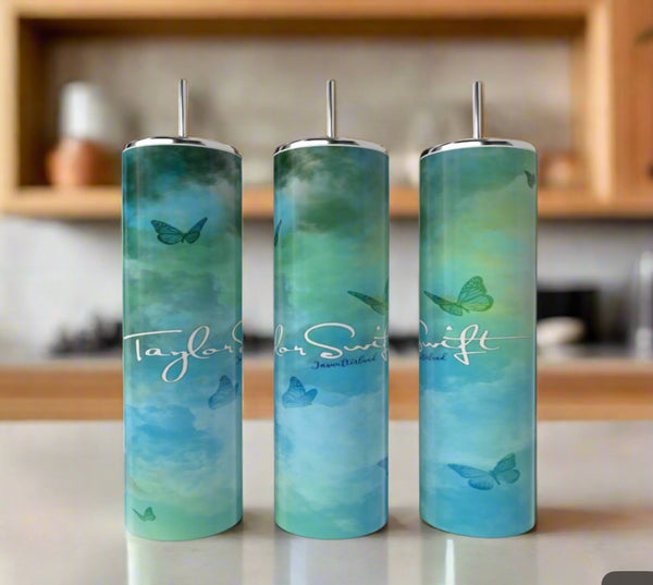 Three Debut Taylor Swift 20oz Tumblers by Kreative Kreationz with metal straws sit on a kitchen counter. The eco-friendly, stainless steel tumblers feature a blue-green gradient, butterfly designs, and white “Taylor Swift” text.