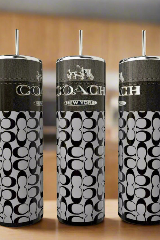 Three Kreative Kreationz Coach Inspired 20oz Skinny Tumblers with straws, featuring a black and white logo and pattern, are displayed on a wooden surface with a blurred background.