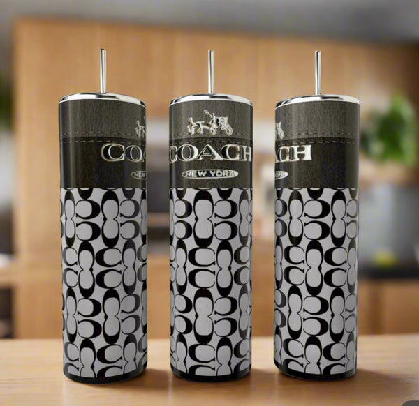 Three Kreative Kreationz Coach Inspired 20oz Skinny Tumblers with straws, featuring a black and white logo and pattern, are displayed on a wooden surface with a blurred background.