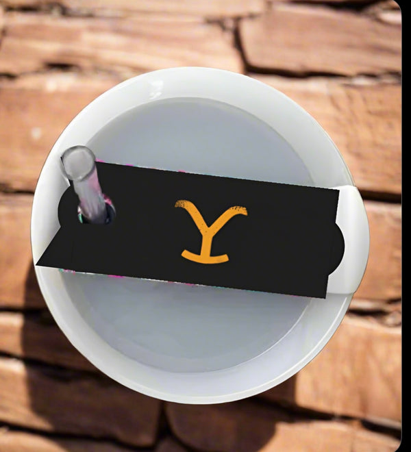 The white Yellowstone Stanley Lid Topper cup by Kreative Kreationz, partially encased in black paper with a yellow "Y" logo, sits on wood. A straw elegantly extends through the lid, hinting at its premium materials and craftsmanship.