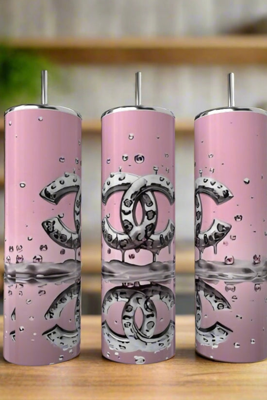 Three 20oz pink Chanel Splash Tumblers by Kreative Kreationz with metallic straws and silver logos sit on a wooden surface, accented by water droplets. This stylish drinkware set adds elegance to any kitchen décor.