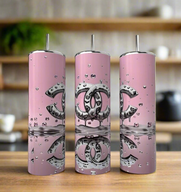 Three 20oz pink Chanel Splash Tumblers by Kreative Kreationz with metallic straws and silver logos sit on a wooden surface, accented by water droplets. This stylish drinkware set adds elegance to any kitchen décor.