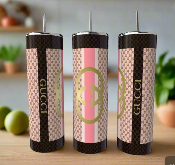 Three Gucci Pink 20oz Skinny Tumblers by Kreative Kreationz, featuring pink, beige, and black Gucci-inspired patterns and logos, are displayed on a wooden surface with blurred kitchen items in the background.