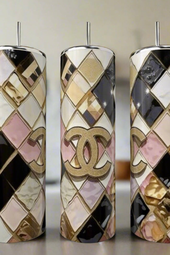 The Chanel Geo 20oz Skinny Tumbler by Kreative Kreationz features a luxury-inspired gold Chanel logo and geometric diamond patterns in gold, pink, black, and white. The image shows three tumblers with metal straws in a blurred kitchen background.