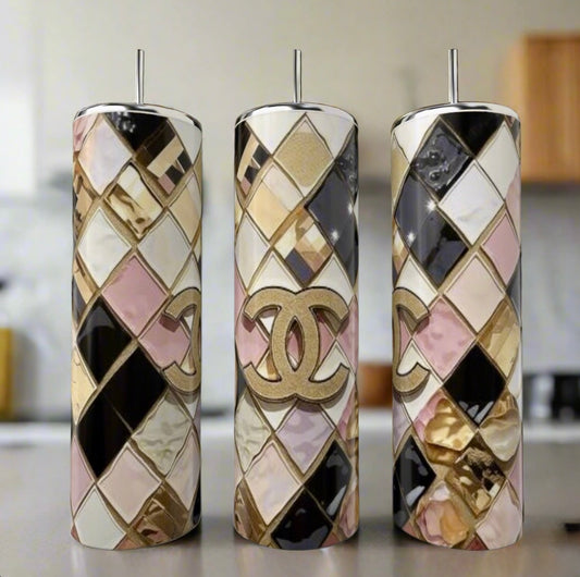 The Chanel Geo 20oz Skinny Tumbler by Kreative Kreationz features a luxury-inspired gold Chanel logo and geometric diamond patterns in gold, pink, black, and white. The image shows three tumblers with metal straws in a blurred kitchen background.