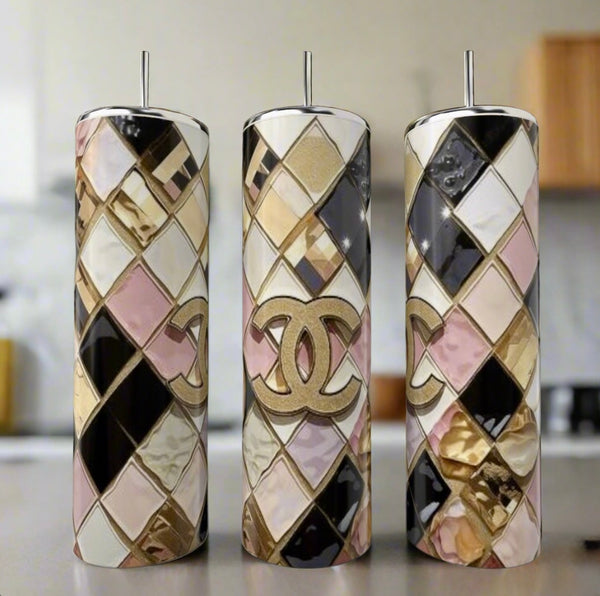 The Chanel Geo 20oz Skinny Tumbler by Kreative Kreationz features a luxury-inspired gold Chanel logo and geometric diamond patterns in gold, pink, black, and white. The image shows three tumblers with metal straws in a blurred kitchen background.