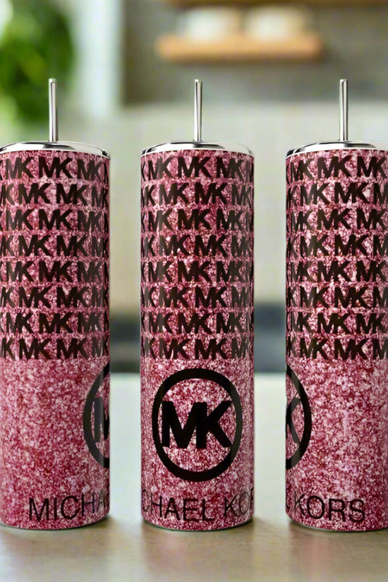Three tall pink glittery Kreative Kreationz Kors Glittered 20oz Tumblers with silver straws are displayed, each featuring an “MK” logo and monogram pattern for a luxe look on a kitchen counter against a blurred background.