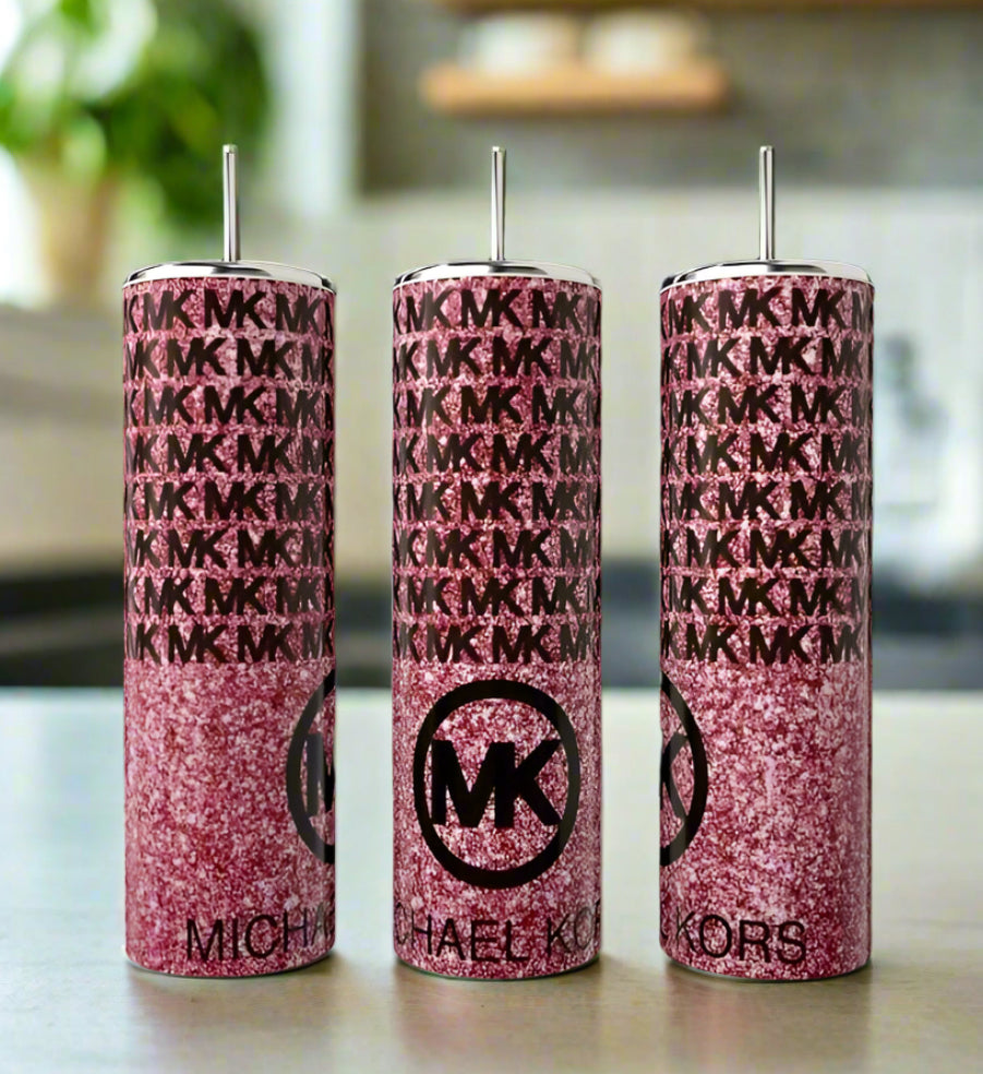 Three tall pink glittery Kreative Kreationz Kors Glittered 20oz Tumblers with silver straws are displayed, each featuring an “MK” logo and monogram pattern for a luxe look on a kitchen counter against a blurred background.