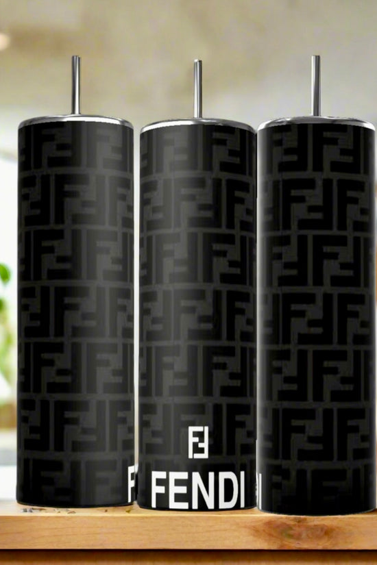Three FENDI Black Grey Modern 20oz Skinny Tumblers by Kreative Kreationz, featuring the FF logo and metal straws, sit on a wooden kitchen surface with blurred plants and fruit in the background. Crafted from high-quality stainless steel.