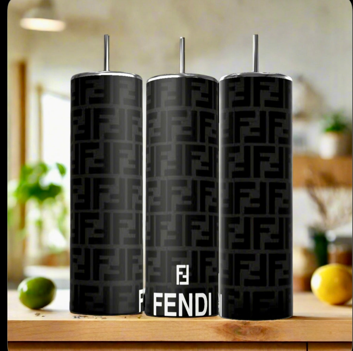 Three FENDI Black Grey Modern 20oz Skinny Tumblers by Kreative Kreationz, featuring the FF logo and metal straws, sit on a wooden kitchen surface with blurred plants and fruit in the background. Crafted from high-quality stainless steel.